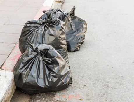 Two large black rubbish bags made of thick plastic are positioned on a paved sidewalk next to a curb, with one bag slightly leaning against the other. The bags contain waste materials, evident from their bulging and irregular shapes. One bag shows a partially twisted top, suggesting it was tightly tied, while the other bag has a crumpled appearance. The curb is constructed from concrete with a red-painted edge, and the paving consists of flat, rectangular stone slabs in varying shades of grey. In the background, the asphalt road surface appears smooth and dark, suggesting an outdoor urban environment, likely near a property in Tottenham, UK. The scene is evenly lit, indicating daytime conditions without harsh shadows, consistent with typical street scenes where rubbish collection or clearance by services such as Office Clearance Tottenham might take place.