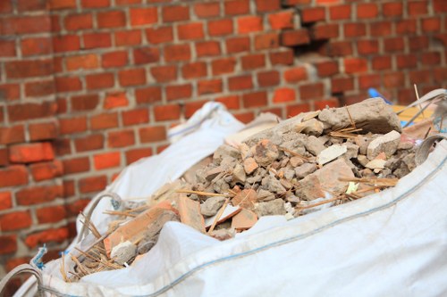 A large white construction rubble sack filled with a mixture of broken bricks, concrete fragments, and assorted debris, situated outdoors on a pavement near a red brick wall. The rubble includes irregularly shaped pieces with rough textures, in shades of tan, light brown, and grey. Some wooden and plastic debris are visible among the rubble. The sack is partially open at the top, allowing the debris to spill slightly over the edges. The background features a red brick wall with a standard running bond pattern, and the area appears to be part of an external property boundary, consistent with rubbish removal or clearance activities in Tottenham. The scene is well-lit, showcasing natural daylight, emphasizing the heap's construction waste materials, consistent with the services offered by Office Clearance Tottenham in removing such refuse.