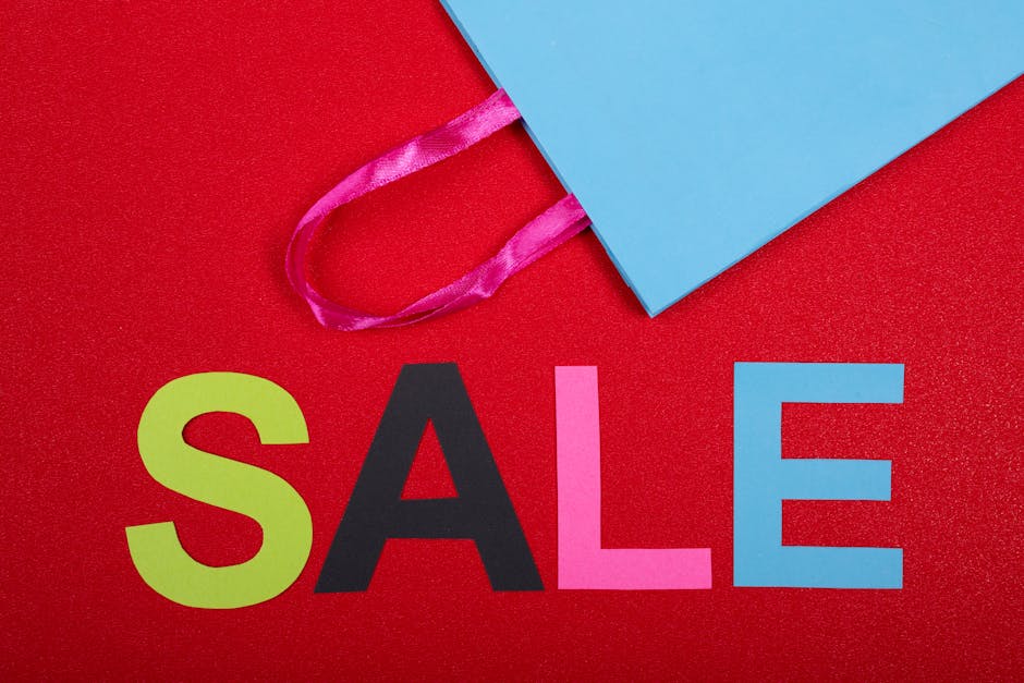 A close-up of a person's left hand holding a white rectangular tag with a pointed end, displaying the word 'SALE' in bold, red glittery letters. The hand is positioned on the left side of the image, w