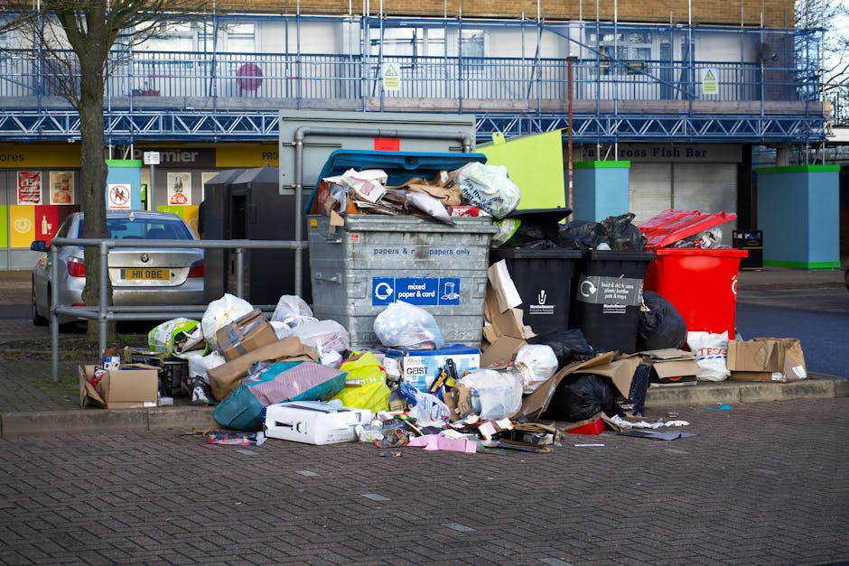 An overflowing set of waste and rubbish bins situated on a paved area in an urban environment, with various types of domestic and commercial waste spilling onto the sidewalk. The central grey bin labe
