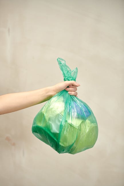 A green waste skip positioned on a paved surface against a dark brick wall, filled with several red and black bags of garden soil or compost, some of which are labeled with brand and product details. 