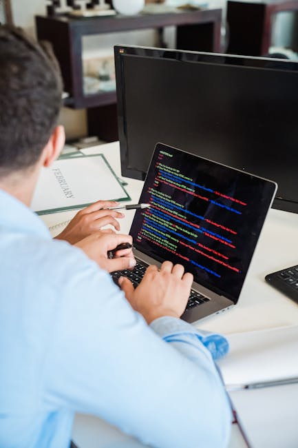 A close-up view of a person's hands typing on a modern laptop with a slim metallic body, placed on a dark, reflective table. The laptop screen displays lines of computer code with syntax highlighting