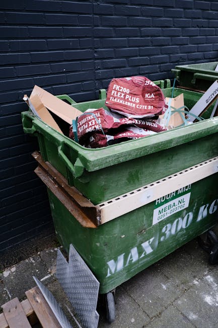 A green wheeled waste skip is positioned on a paved surface outdoors against a dark brick wall. The skip contains several red bags labeled with white text, indicating they are from a brand of flexible